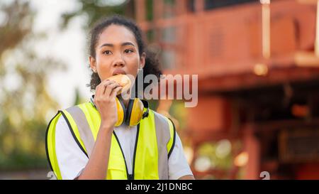 smile African american woman foreman worker  or woman maintenance engineer in reflective vest safety jacket sits down on old truck for relaxing, eats Stock Photo