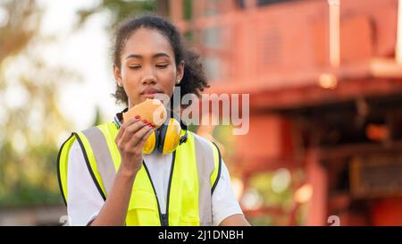 smile African american woman foreman worker  or woman maintenance engineer in reflective vest safety jacket sits down on old truck for relaxing, eats Stock Photo