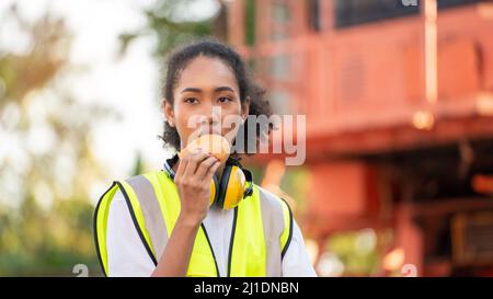 smile African american woman foreman worker  or woman maintenance engineer in reflective vest safety jacket sits down on old truck for relaxing, eats Stock Photo