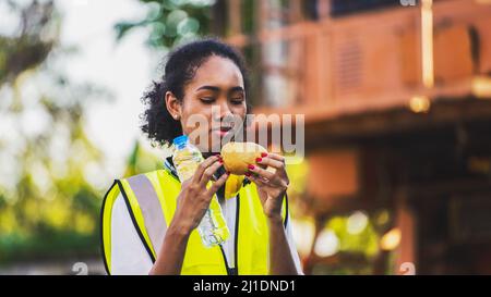 smile African american woman foreman worker  or woman maintenance engineer in reflective vest safety jacket sits down on old truck for relaxing, eats Stock Photo