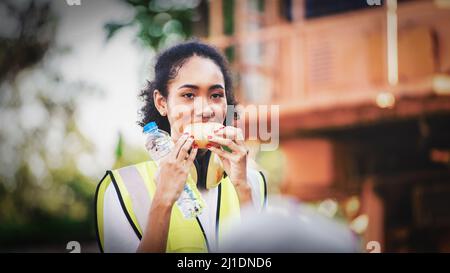 smile African american woman foreman worker  or woman maintenance engineer in reflective vest safety jacket sits down on old truck for relaxing, eats Stock Photo