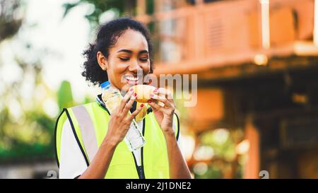 smile African american woman foreman worker  or woman maintenance engineer in reflective vest safety jacket sits down on old truck for relaxing, eats Stock Photo