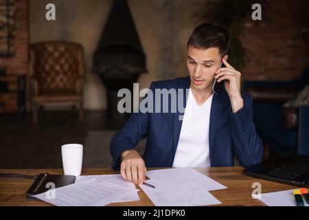 Young businessman is sitting at table working with documents talking on mobile phone Documents on laptop table. Freelancer works from home. Telephone conversations. Job certification online education Stock Photo