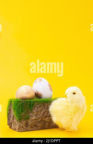 Chicken next to stone pedestal with grass and painted gold and white eggs on yellow background. Easter. Copy space Stock Photo