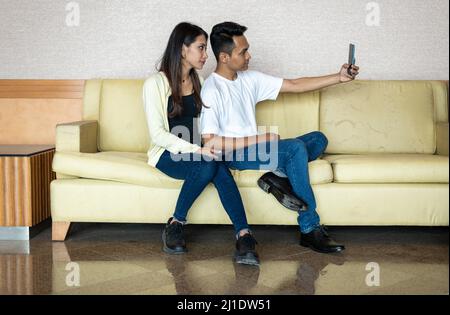 Malay Asian Malaysian Muslim Couple happy taking a selfie photo using a hand phone, while sitting on a light brown settee sofa chair at home Stock Photo