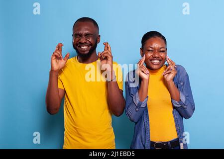 Hopeful people crossing fingers and hoping for good luck in studio. Enthusiastic couple praying for miracle sign and having positive expectations, feeling superstitious about belief. Stock Photo