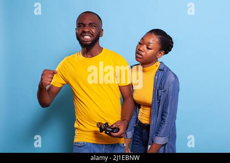 Frustrated couple losing video games on console with joystick in studio. Disappointed boyfriend and girlfriend using controller to play online game, feeling sad about losing in front of camera. Stock Photo