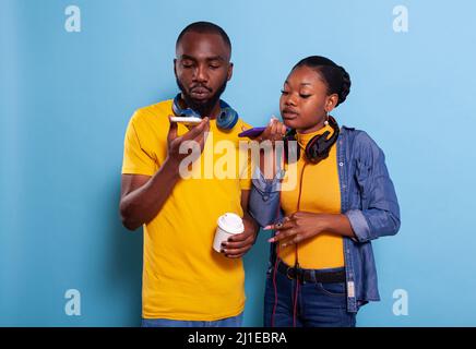 Modern couple using speaker on smartphone to record message, wearing headset over blue background. Boyfriend and girlfriend holding mobile phone to use microphone, making speech online. Stock Photo