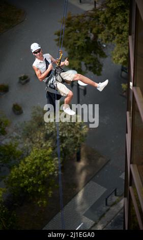Industrial mountaineering worker in protective helmet showing approval gesture and smiling while hanging on climbing rope outside building. Man in sunglasses using safety lifting equipment outdoors. Stock Photo