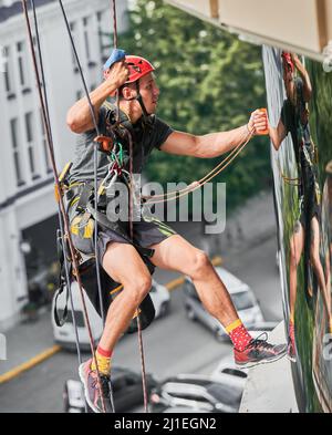 Industrial mountaineering professional cleaner hanging on safety climbing rope and washing building facade. Man cleaning service worker using safety lifting equipment while wiping window outdoors. Stock Photo