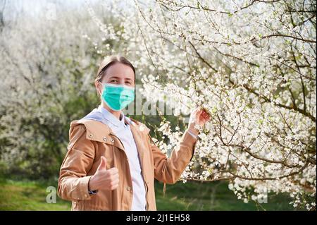 Woman allergic suffering from seasonal allergy at spring, posing in blossoming garden at springtime. Happy woman wearing medical mask near blooming trees, showing thumbs up. Spring allergy concept Stock Photo