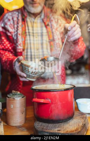 man in casual rustic clothes pours soup from large authentic pot into ceramic plate with ladle Stock Photo