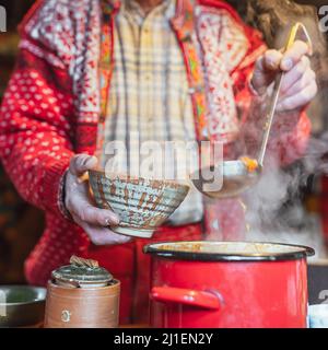 man in casual rustic clothes pours soup from large authentic pot into ceramic plate with ladle Stock Photo