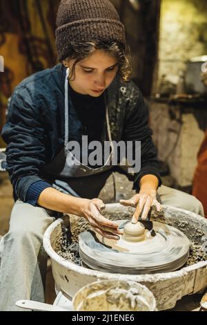 teenage girl in working clothes stained with clay is studying pottery, learning to work on potter's wheel in potter's workshop. Stock Photo