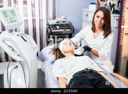 Skilled female beautician making patient non-surgical ultrasound SMAS lifting, only non-invasive procedure that tightening skin of neck, chin and eyebrows in special clinic of beauty. Stock Photo