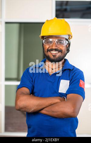 construction worker with safety helmet and eye glasses standing with crossed arms by looking at camera at site - concept of blue collar jobs Stock Photo