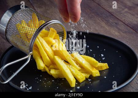 Still life with a portion of freshly made crispy french fries with a bright yellow colour on a black plate and a person's fingers sprinkling salt on t Stock Photo