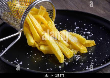 Still life with a portion of freshly made crispy french fries with a bright yellow colour on a black plate with salt on top. Stock Photo