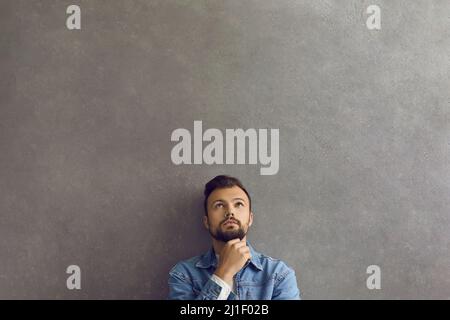 Handsome young man leaning on grey wall, thinking and looking up at copy space above Stock Photo