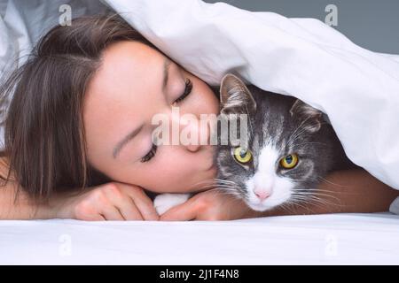 Lovely woman kissing her beautiful cat under the white blanket in the bed. Cat and owner together. Love for cats Stock Photo