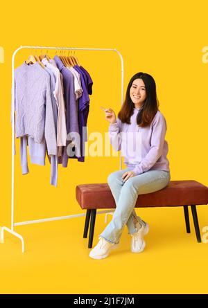 Young Asian woman pointing at rack with clothes in purple shades on yellow background Stock Photo