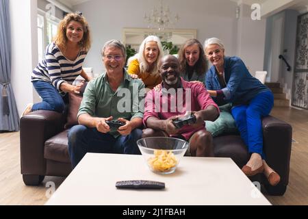 Multiracial senior male friends playing video game while women cheering them in background at home Stock Photo