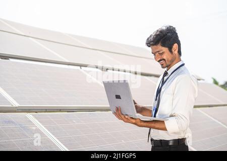 smiling engineer checking operation by working on laptop in front of solar panel at farmland - concept of technology, sustainable lifestyle and Stock Photo