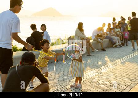 15 10 2021 boy and girl, parents have fun, blowing soap bubbles in West Kowloon Waterfront Promenade, Hong Kong in evening. backlight shot Stock Photo