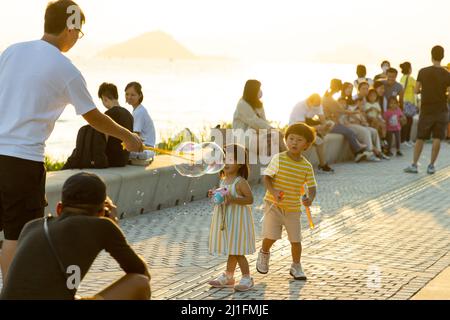 boy and girl, parents have fun, blowing soap bubbles in West Kowloon Waterfront Promenade, Hong Kong in evening. backlight shot Stock Photo