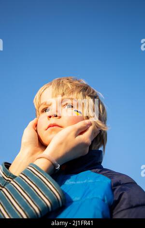portrait of a boy from Ukraine with a painted yellow-blue heart on his cheek and female hands gently hug his face. sadness of little peaceful Ukrainia Stock Photo