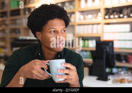 Thoughtful young african american male customer holding coffee mug looking away in coffee shop. unaltered, cafe culture, people and lifestyle concept. Stock Photo