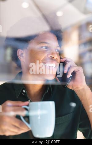 Smiling young african american man talking on smartphone holding coffee mug seen through cafe window. unaltered, cafe culture, people, technology and Stock Photo