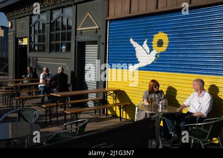 Glasgow, UK, 25th March 2022. A couple enjoy the sun in front of a mural on the wall of BiddyÕs Tea Room depicting the Ukrainian flag and a dove of peace, painted to show support for Ukraine in their ongoing war against the invasion by Russia, in the east end of Glasgow, Scotland, 25 March 2022. Photo credit: Jeremy Sutton-Hibbert/Alamy Live News. Stock Photo