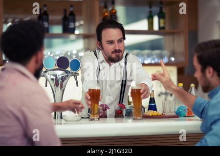 To quench your thirst. Shot of a friendly barman serving beer to two customers at his bar. Stock Photo