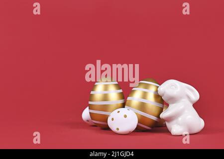 Small Easter bunny figure and golden and white easter eggs with stripes and dots on dark red background with copy space Stock Photo