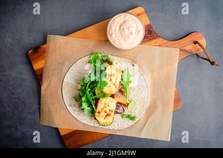 Assembling a Roasted Vegetable and Halloumi Wrap: A whole wheat flatbread topped with roasted vegetables, hummus, halloumi cheese, and harissa sauce Stock Photo