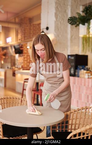 Vertical medium long portrait of young female waitperson wearing mask preparing table cleaning it in cozy cafe Stock Photo