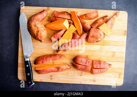 Cutting Unpeeled Sweet Potatoes into Wedges: Cutting up sweet potatoes on a bamboo wood cutting board Stock Photo