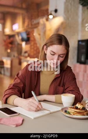 Portrait of attractive young woman sitting at table in modern cafe making notes in notebook during lunch break Stock Photo