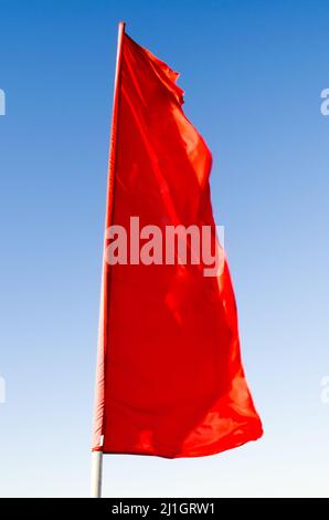 Red flag of rectangular shape elongated fluttering in the wind on the flagpole background of the blue sky Stock Photo