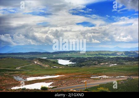 Scenic panorama of Beartooth Pass, in Wyoming, shows cars traversing mountain pass, snow, and cloudscape over blue sky. Stock Photo
