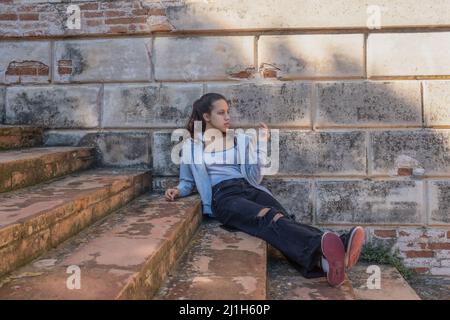 Young woman sits on grey stairs dressed in ripped jeans brick wall behind Stock Photo