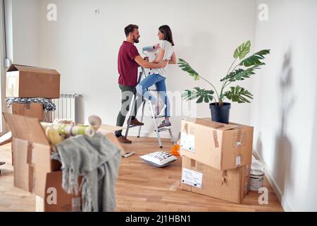 caucasian couple in love having fun while preparing to paint white wall in apartment, standing on a ladder, holding paintbrushes, smiling, laughing, l Stock Photo