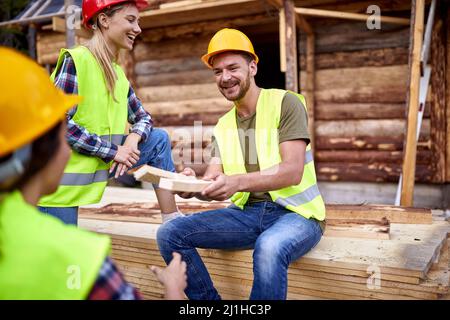 Man giving piece of wooden board over to another construction worker. Smiling construction workers working on wooden house skeleton frame. Constructio