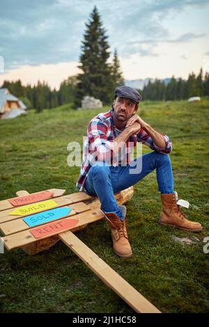 Young man sitting on wood with wooden board beside him. Thinking. Looking into distance.  Wooden Skeleton Framing Building. Construction industry, rea Stock Photo