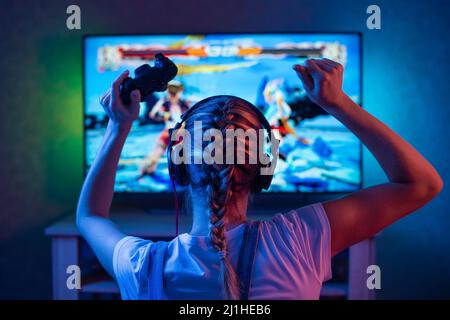 A gamer or a streamer girl at home in a dark room with a gamepad playing with friends on the networks in video games. A young man sits in front of a m Stock Photo