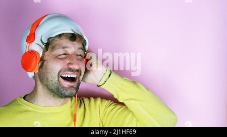 Horizontal studio shot of cheerful carefree bearded man wears white hat, yellow shirt, listening to music with high volume in stereo orange headphones Stock Photo