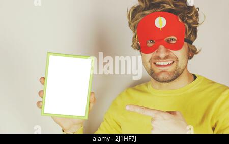 Funny excited young male in red superhero mask looking at camera and pointing at blank picture space on white background . Stock Photo