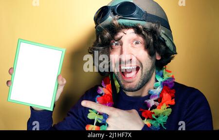 Studio Shop of a funny young man while partying wearing a helmet and a Hawaiian necklace showing blank placard with copy space for your text, yellow b Stock Photo