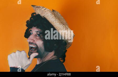 Laughing cheerful young bearded man wearing casual farmer hat pointing thumb aside on mock up copy space isolated or dark orange color background stud Stock Photo